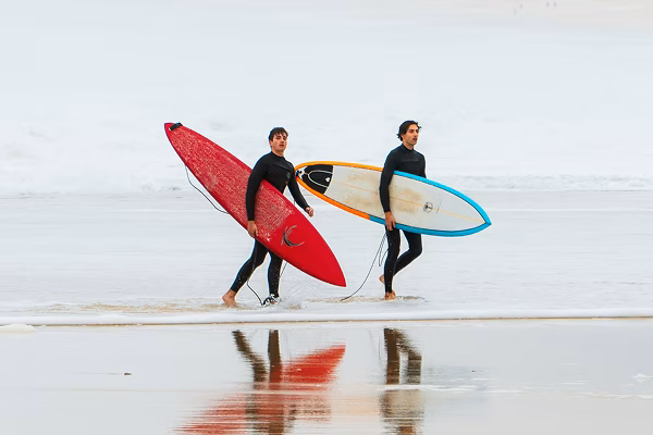 Two surfers in wetsuits walking along a beach with surfboards